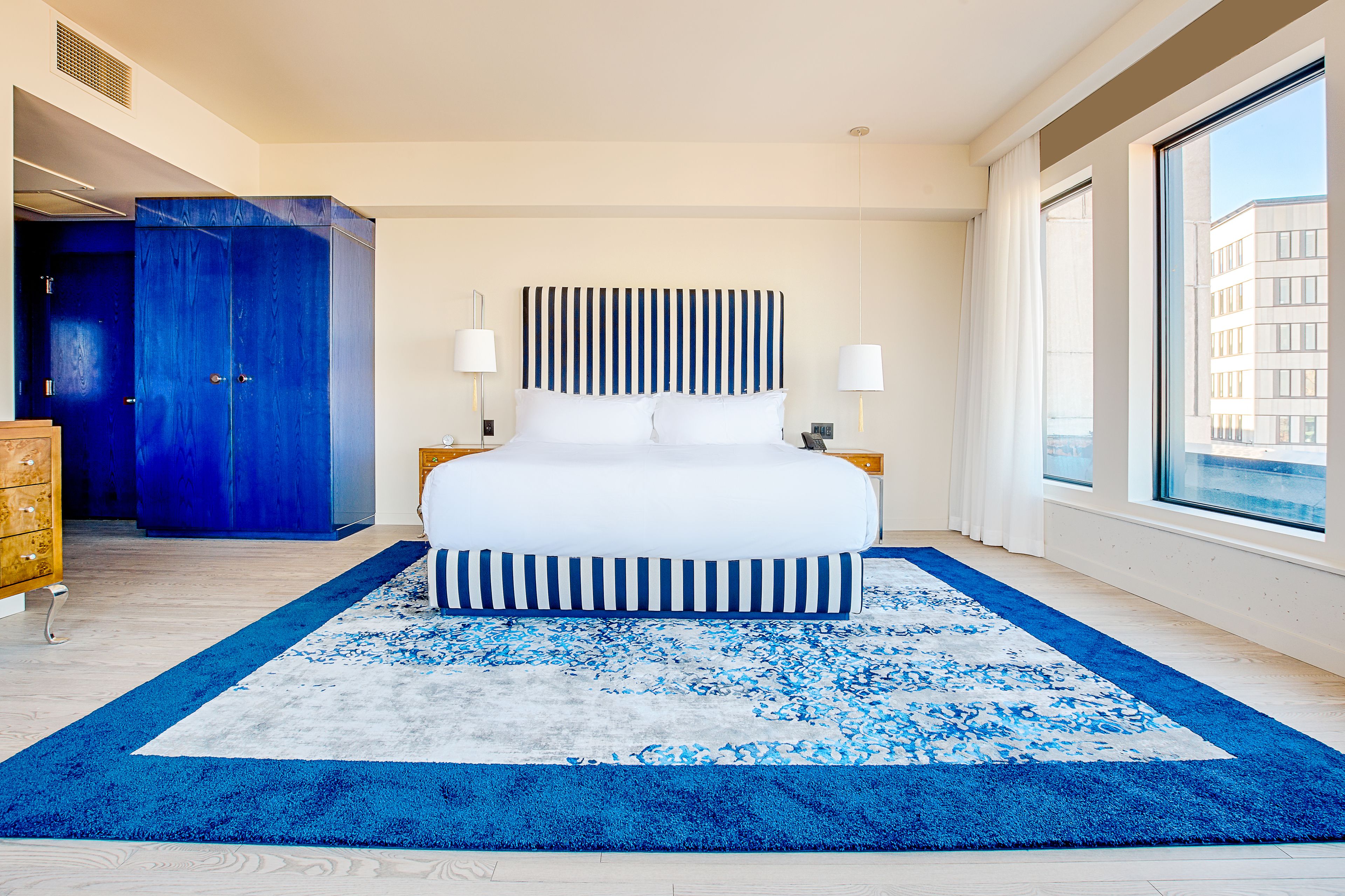Een moderne hotelkamer met een groot bed, een blauw-wit gestreept tapijt en uitzicht op de stad door grote ramen.