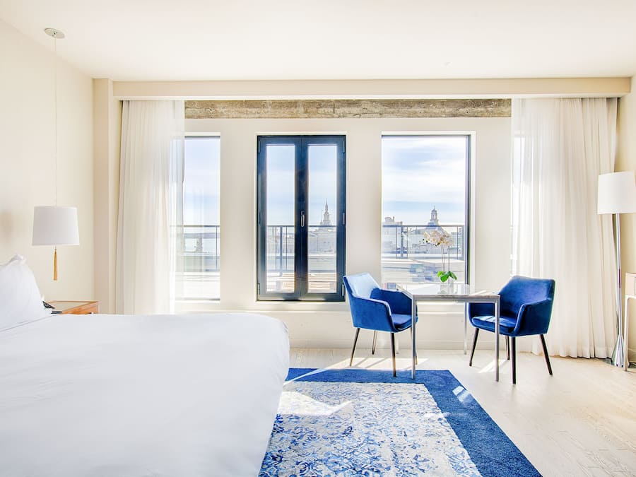 Habitación de hotel con una cama grande, un escritorio con dos sillas, vistas a un paisaje urbano a través de amplios ventanales y una alfombra azul estampada.