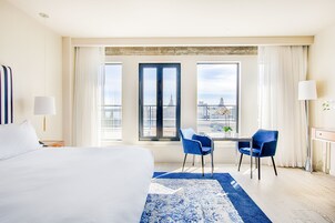 A hotel room with a large bed, a desk with two chairs, a view of a cityscape through large windows, and a blue patterned rug.
