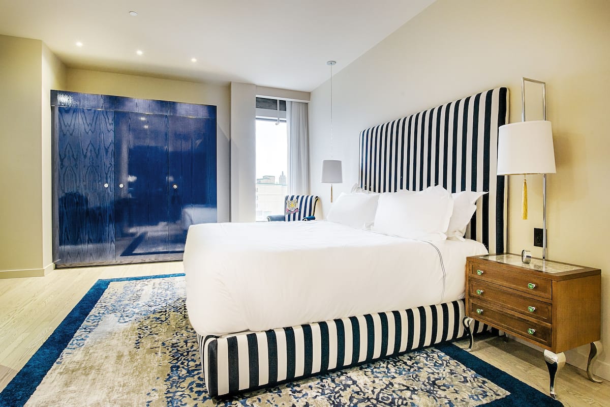 A hotel room with a large bed, a nightstand with drawers, a lamp, and a view of the city through the window.
