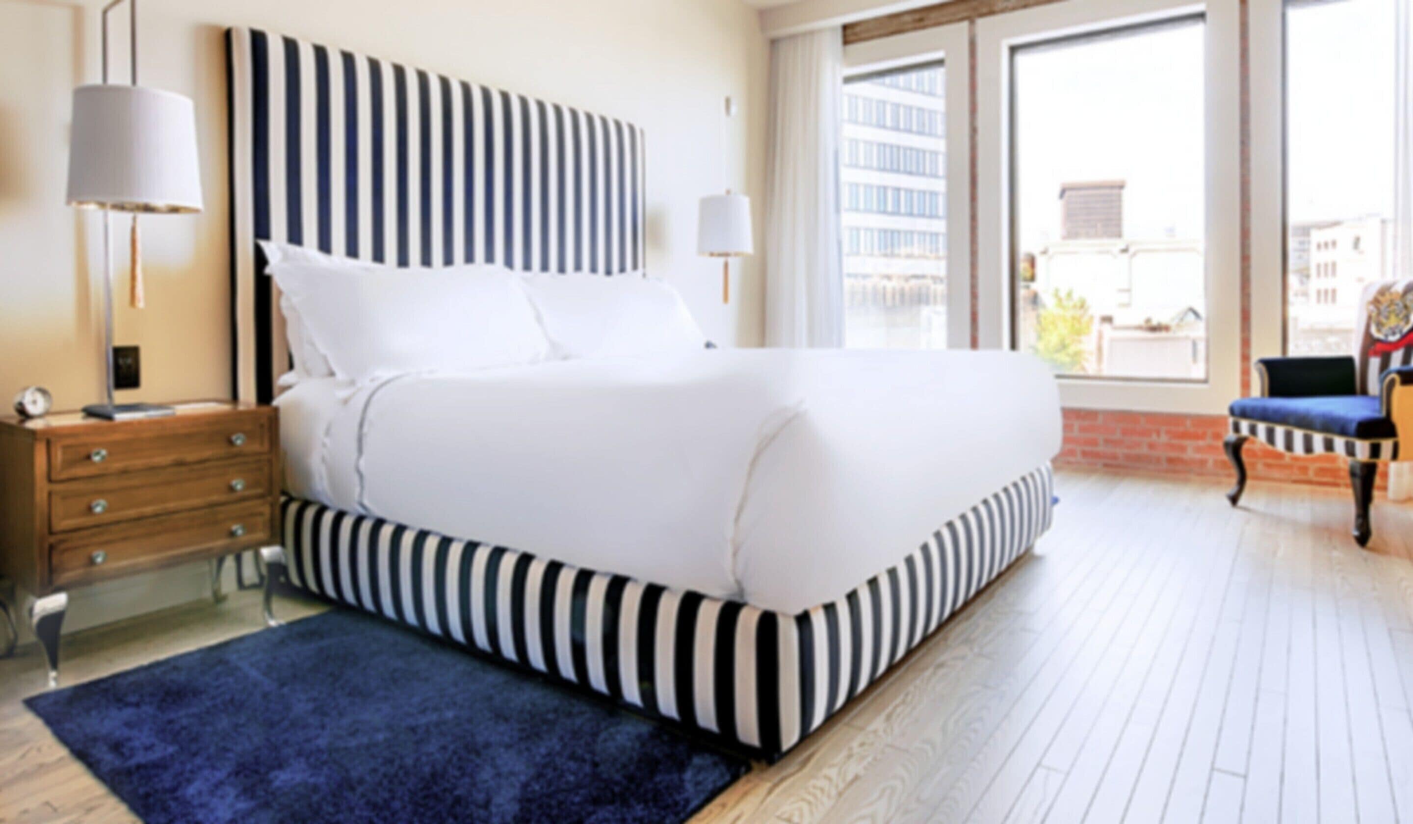 Habitación de hotel con una cama grande, una mesita de noche de madera, un tapete azul y vistas a la ciudad a través de la ventana.