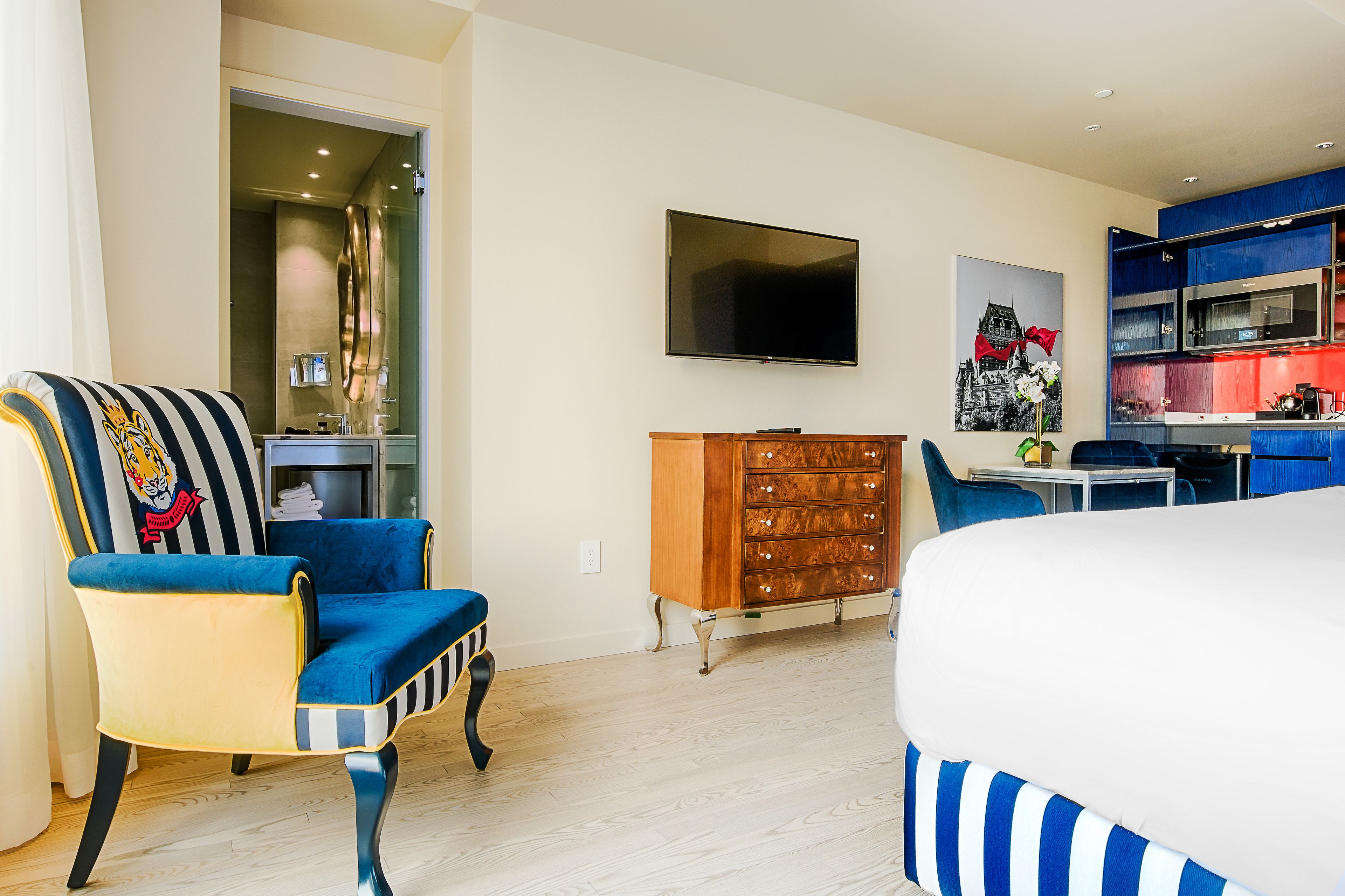 Una camera d'albergo moderna con un letto grande, una TV fissata al muro, un comò in legno e una poltrona a righe blu e bianche.