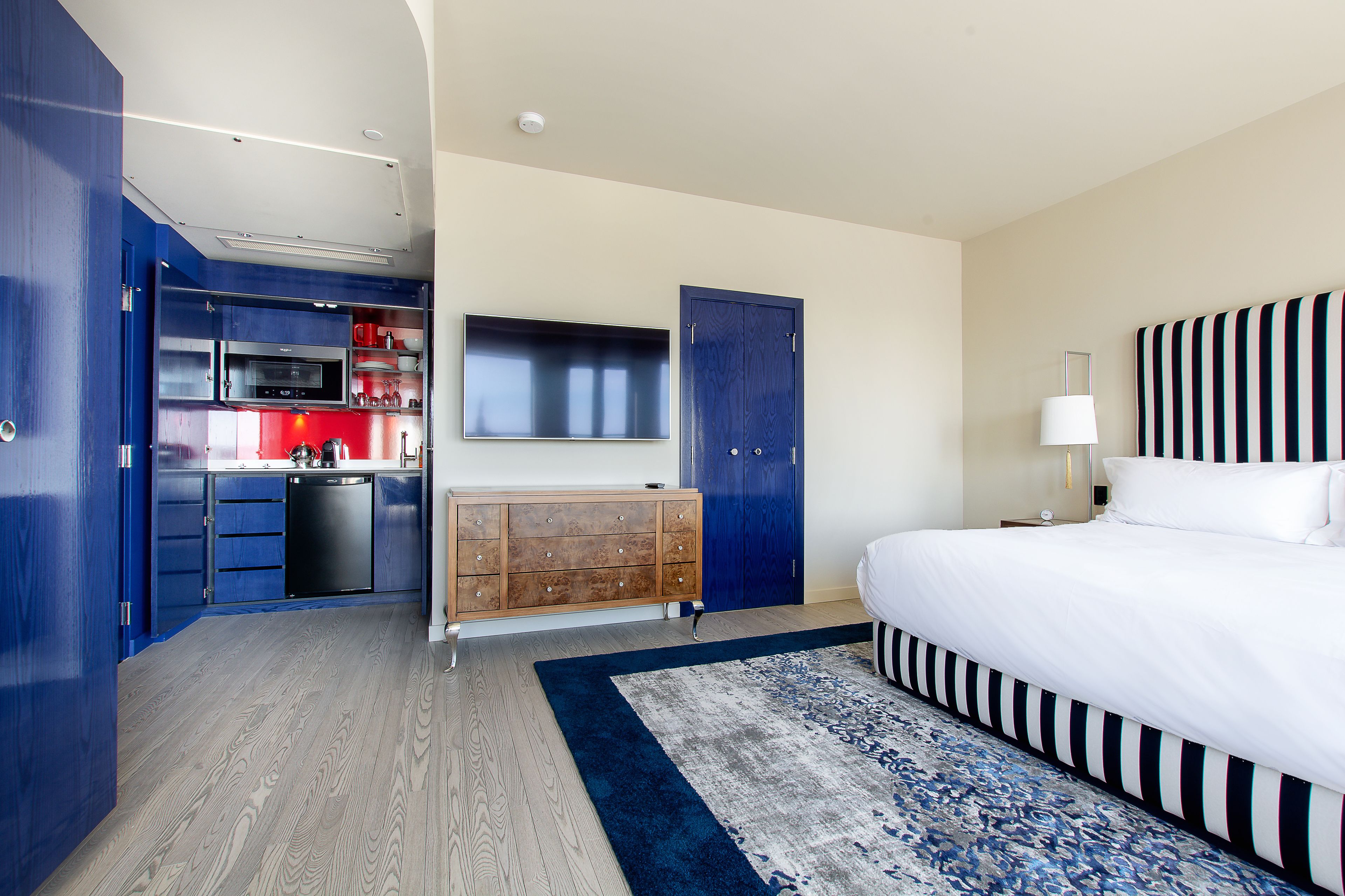Une chambre d’hôtel avec un grand lit, un téléviseur à écran plat fixé au-dessus d’une commode en bois, un coin cuisine avec des placards bleus et une tête de lit bleue et blanche à rayures.