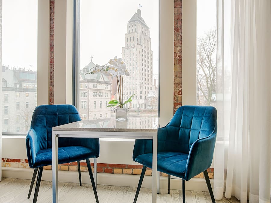 A modern room with two blue velvet chairs, a glass table, and a view of a cityscape through large windows.