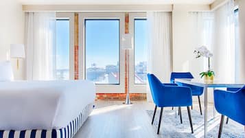 A hotel room with a large bed, a dining table with blue chairs, and a view of the city through large windows.