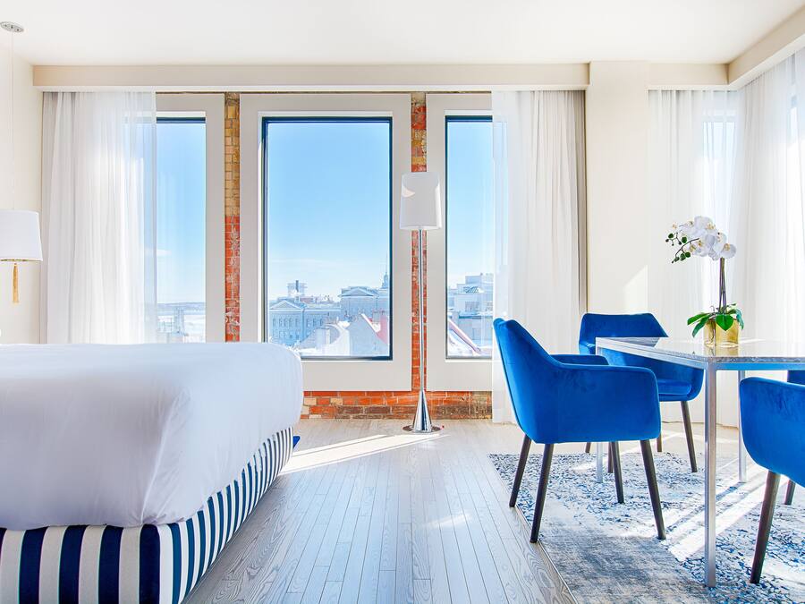 A hotel room with a large bed, a dining table with blue chairs, and a view of the city through large windows.