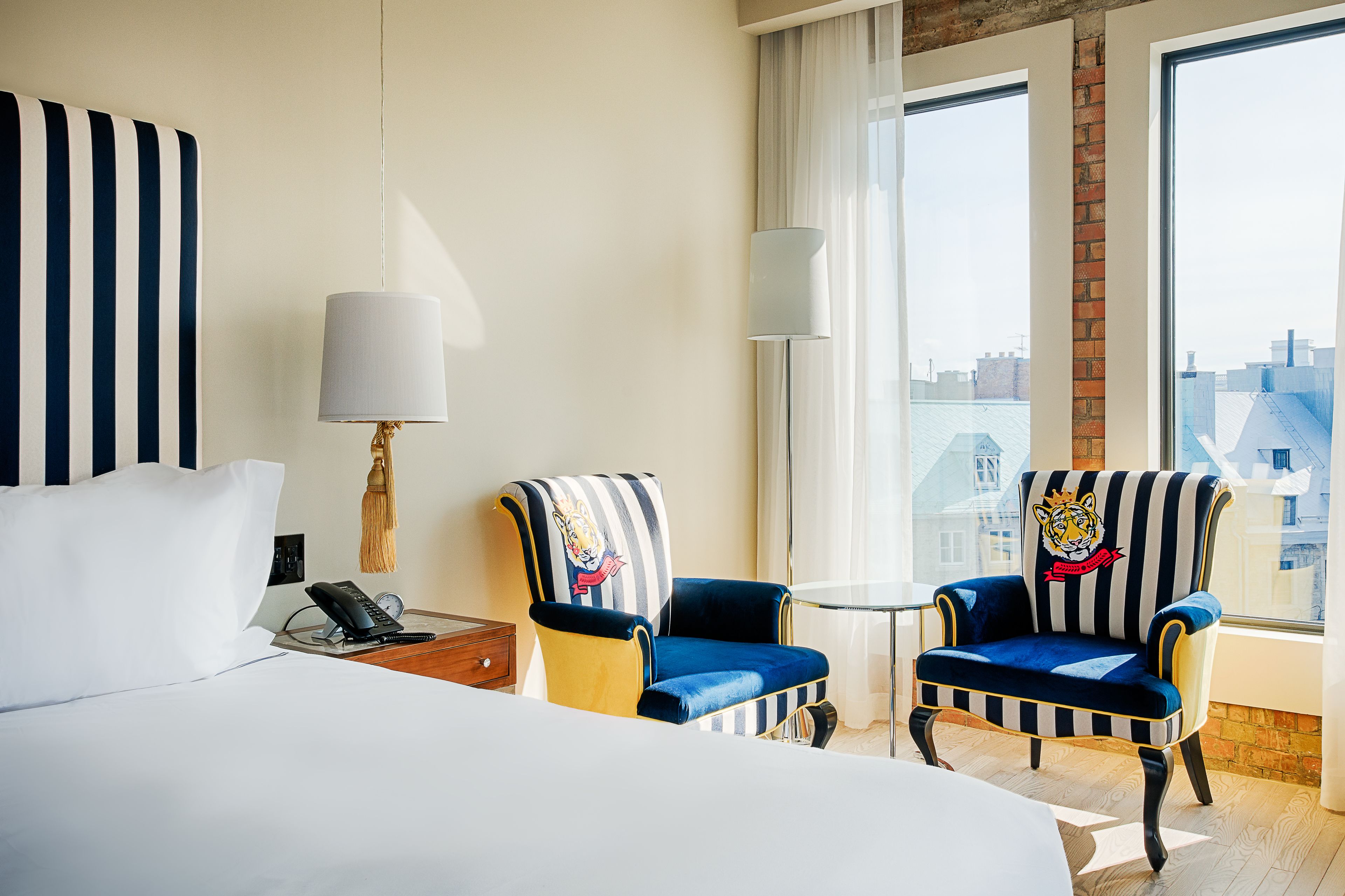 Quarto de hotel com uma cama grande, duas poltronas listradas, uma mesinha, uma luminária e vista para a cidade pela janela.