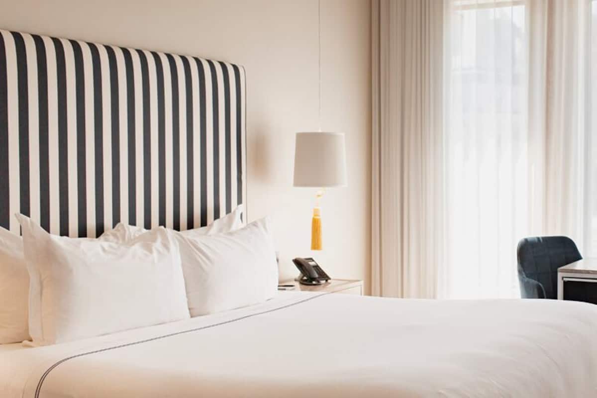A hotel room with a large bed, a bedside table with a lamp, a desk with two chairs, and a window with sheer curtains.