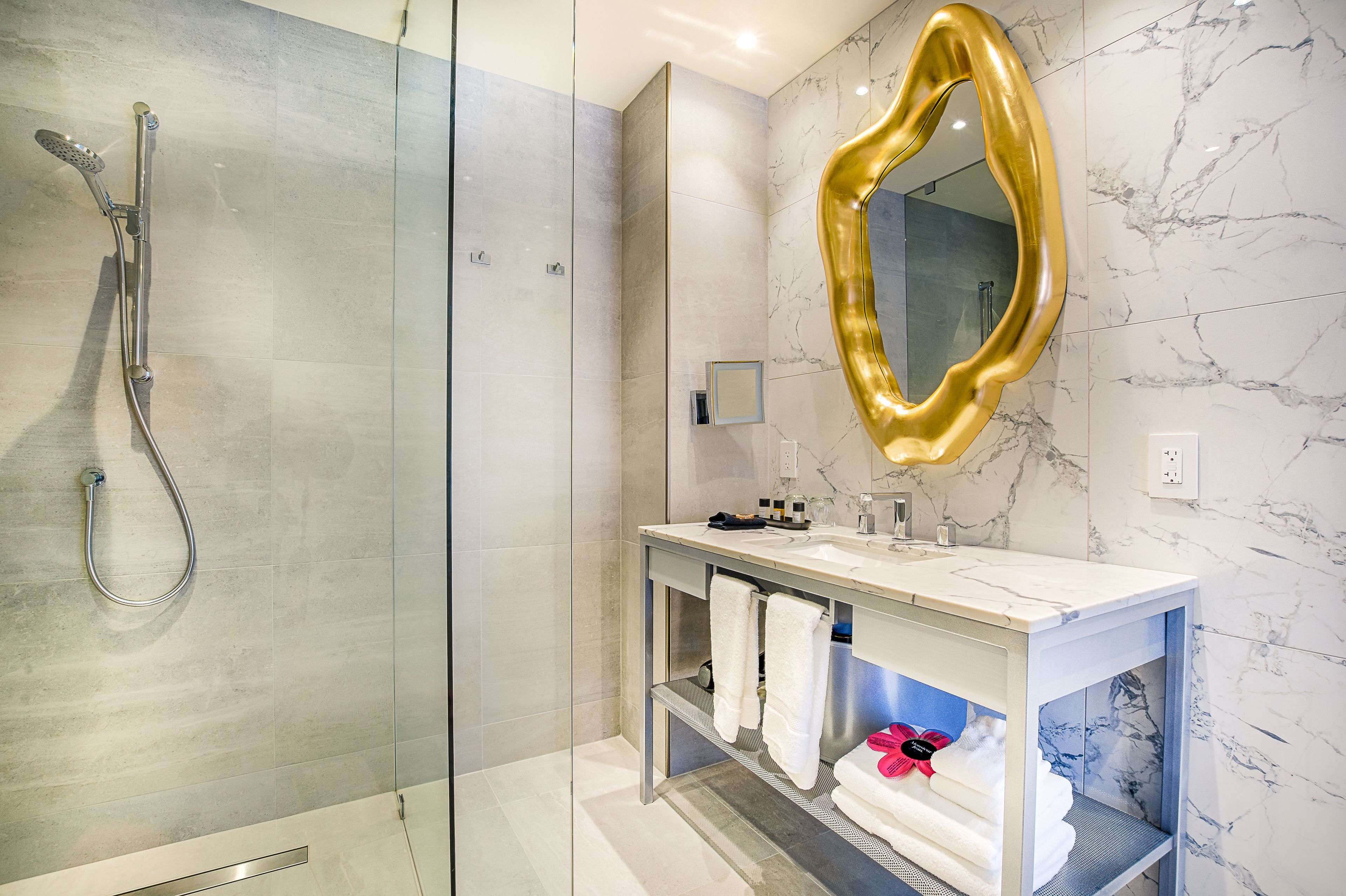 Un baño moderno con ducha de vidrio, pared con azulejos de mármol, espejo con marco dorado y tocador blanco con toallas.