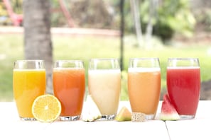 Five glasses of different colored smoothies with slices of fruit in front of a blurred outdoor background. - Bambu Resort (Mazatenango)