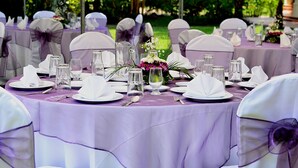 Mesas redondas con sillas blancas y manteles morados, preparadas para un evento formal.