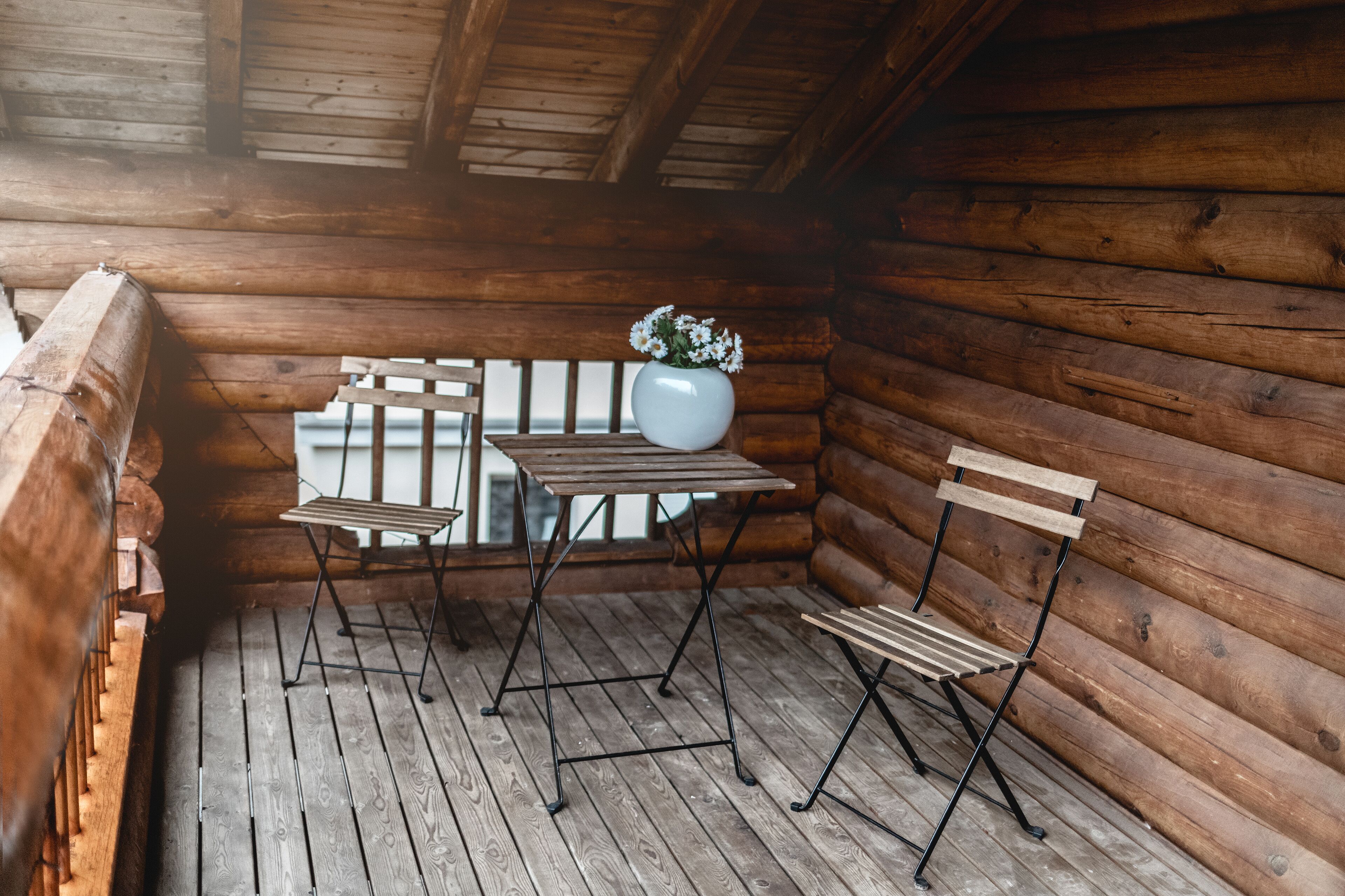 A wooden cabin interior with a wooden table and bench, a white vase with flowers, and a wooden railing.