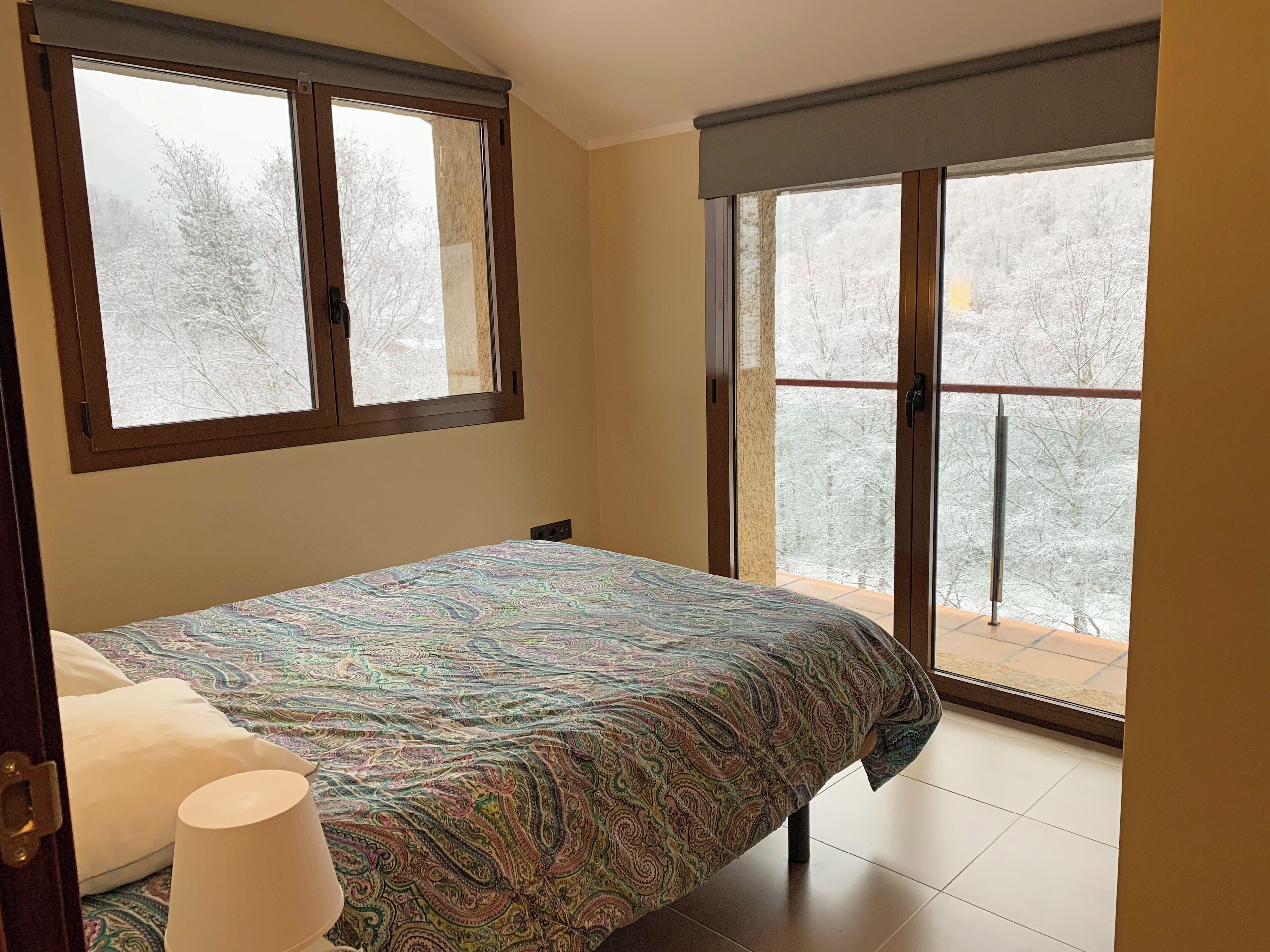 Un dormitorio con cama, ventana y balcón con vistas a árboles nevados.