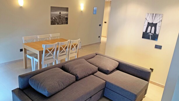 A modern living room with a grey sectional sofa, a wooden dining table with white chairs, and two wall-mounted light fixtures. - Apartamentos Llorts Ordino 3000 (La Cortinada)