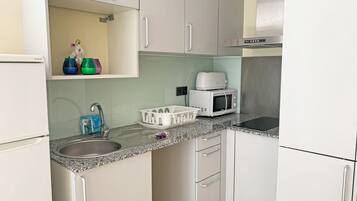 Cocina compacta con un lavamanos, microondas y tostadora sobre la encimera. Hay armarios con tiradores y un estante pequeño con objetos decorativos.