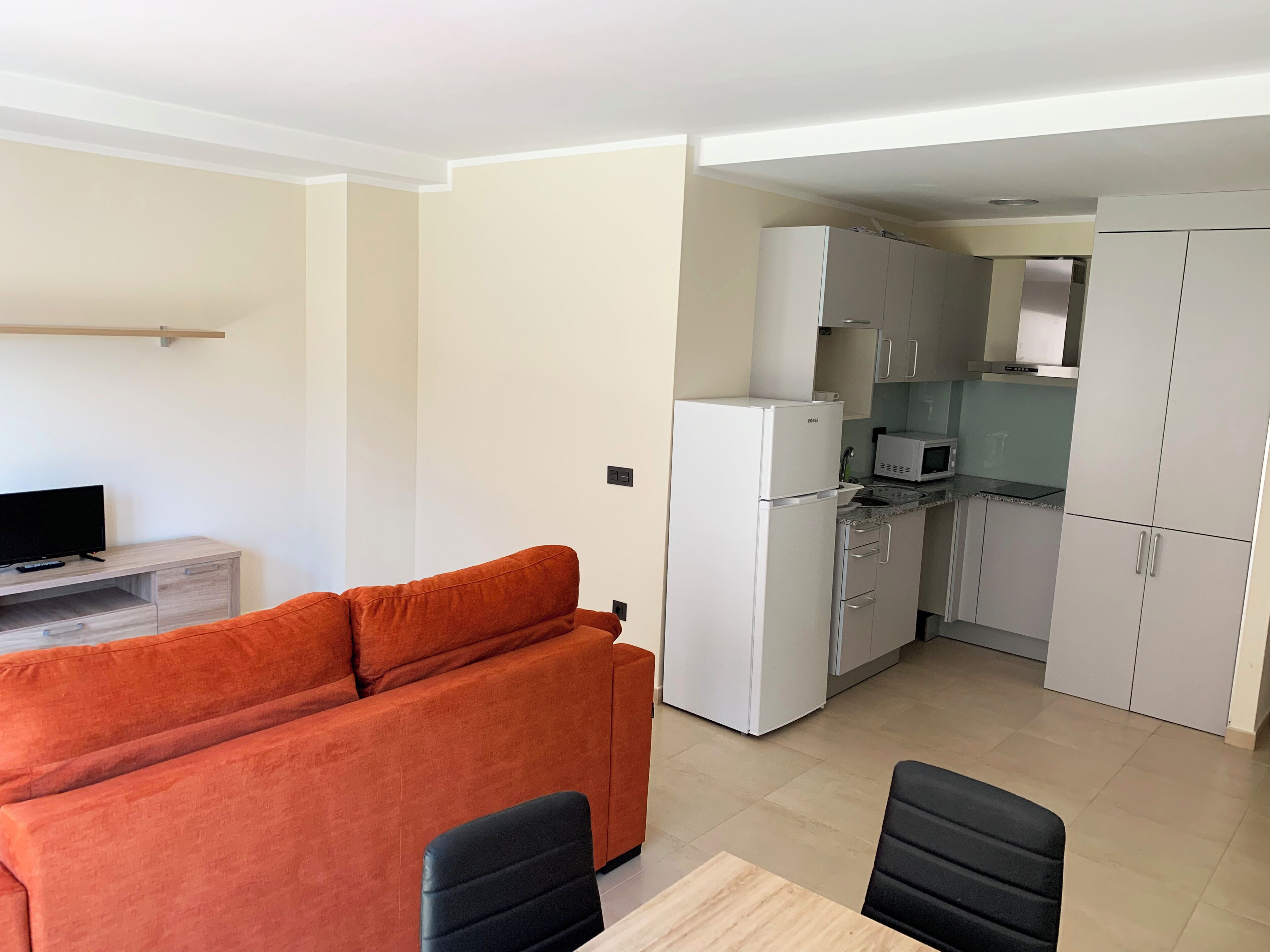 A compact living space with a sofa, a dining table with chairs, a kitchenette with a refrigerator and microwave, and a television on a stand.