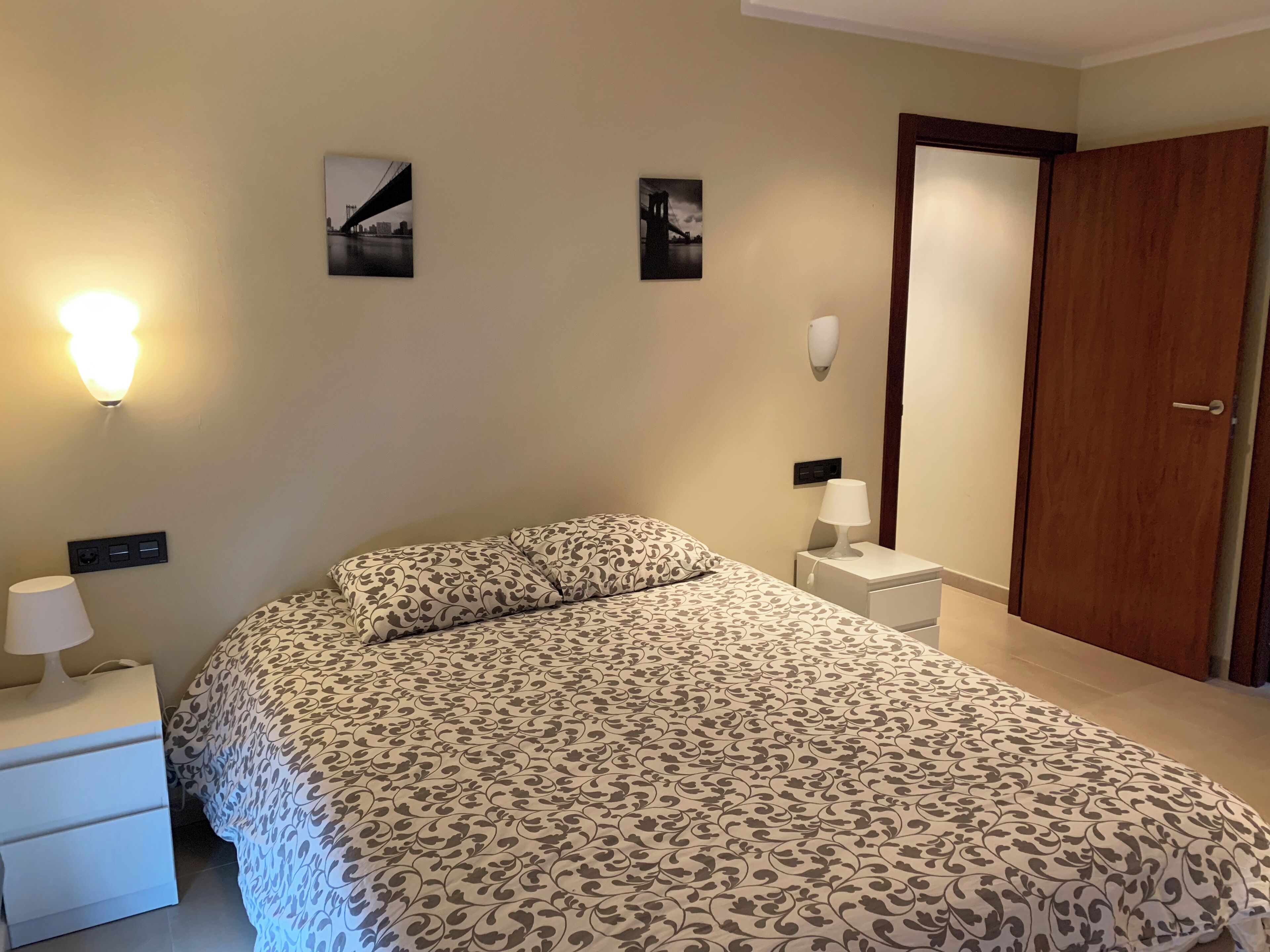 A bedroom with a bed, two bedside tables, a nightstand, a wall-mounted lamp, and a door.