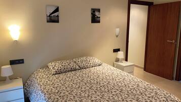 A bedroom with a bed, two bedside tables, a nightstand, a wall-mounted lamp, and a door.