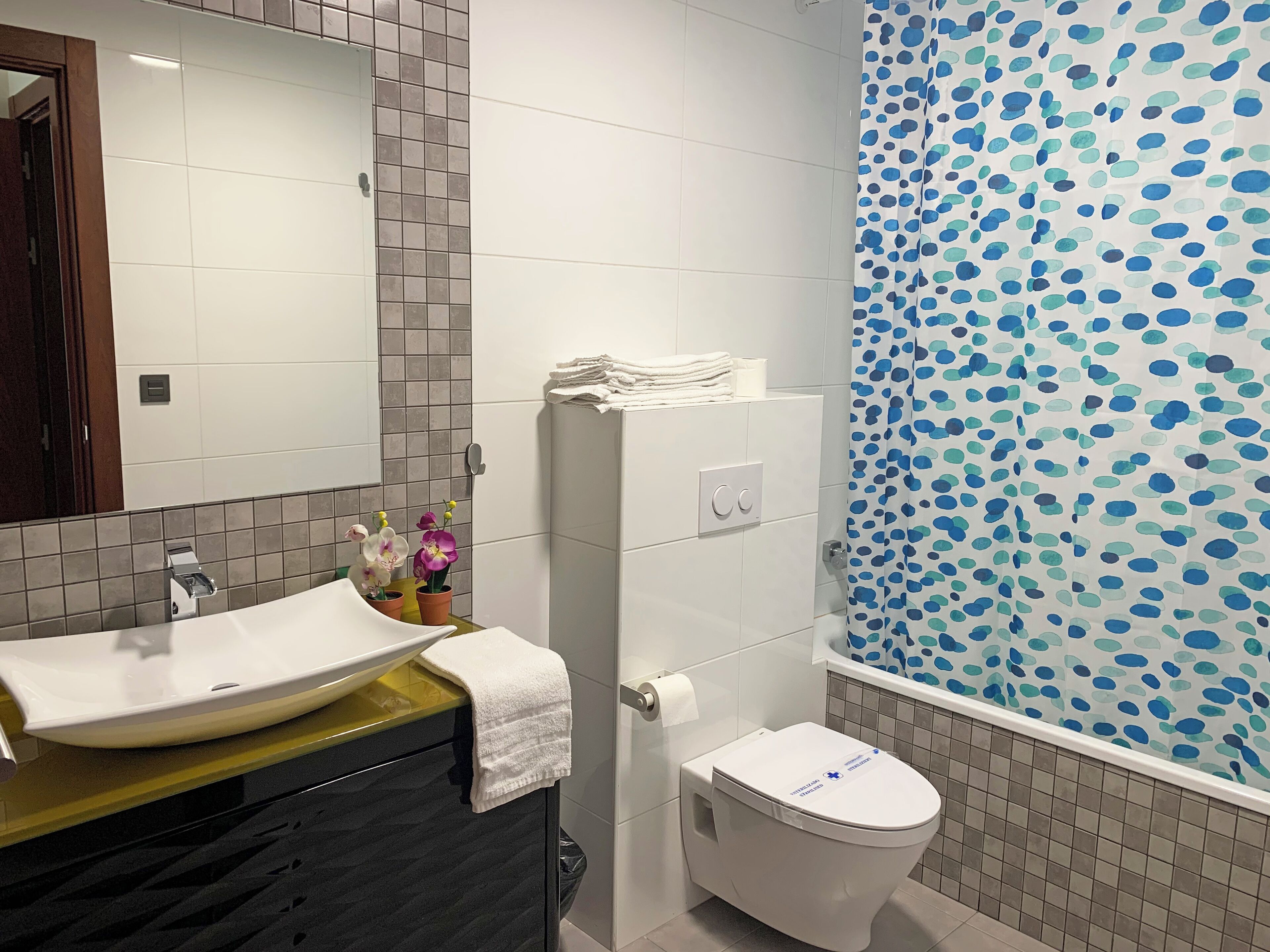 Un baño con lavamanos, espejo y retrete blancos. Hay toallas sobre el lavamanos y una cortina de ducha con diseño azul.