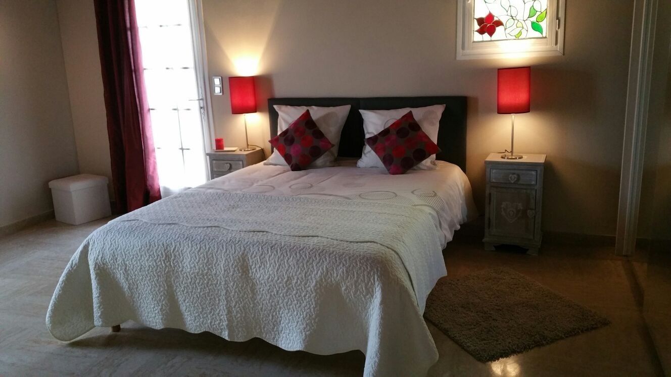A neatly made bed with a white duvet, two red and black patterned pillows, and a white headboard. There are two bedside tables with lamps, a small white trash bin, and a red curtain partially drawn.