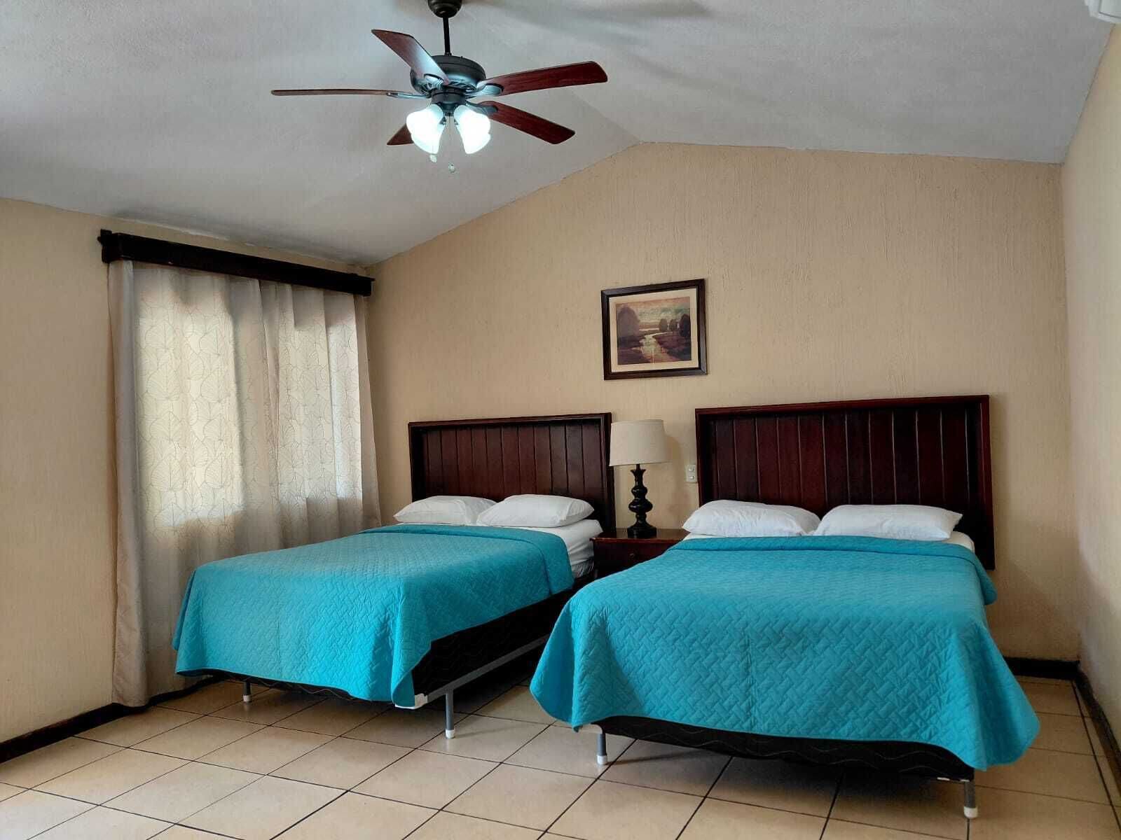 Habitación de hotel con dos camas individuales, un ventilador de techo, una mesita de noche con lámpara y un cuadro en la pared.