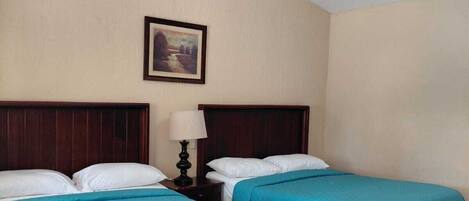 A hotel room with two single beds, a ceiling fan, a nightstand with a lamp, and a framed picture on the wall.