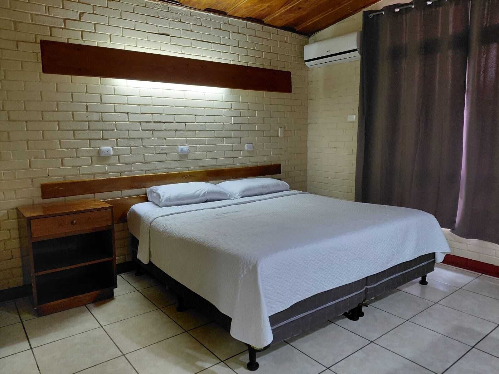 A bedroom with a brick wall, a wooden headboard, a bed with white bedding, a nightstand, and a window with a curtain.