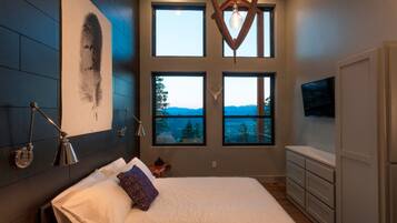 Un dormitorio con una cama grande, varias ventanas, un televisor montado y una lámpara de pared.