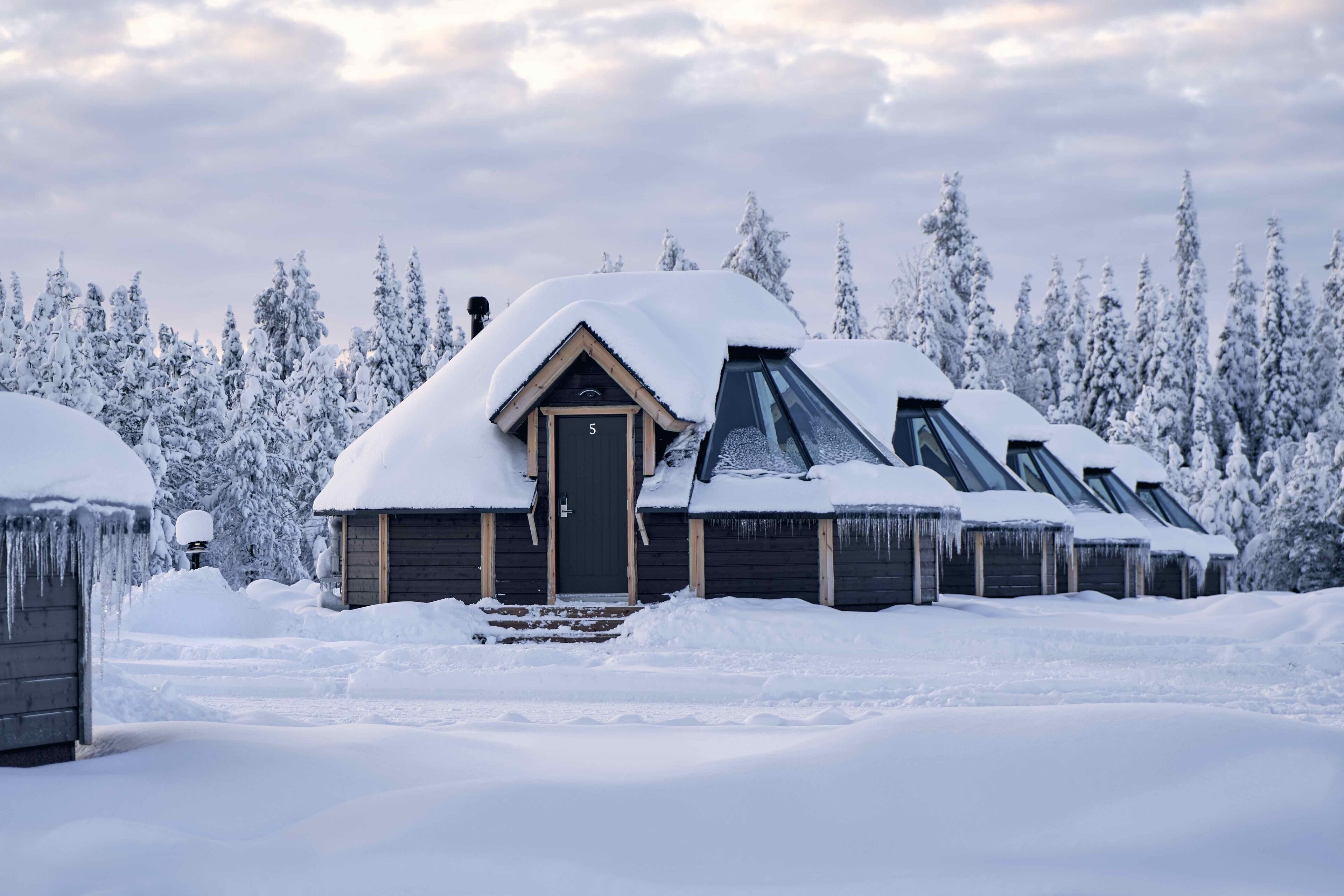 Une rangée de chalets en rondins, aux toits recouverts de neige et dotés d’une porte en bois, s’étend au milieu d’un paysage enneigé.