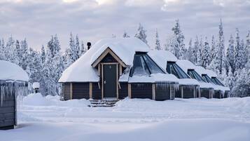 Une rangée de chalets en rondins, aux toits recouverts de neige et dotés d’une porte en bois, s’étend au milieu d’un paysage enneigé.