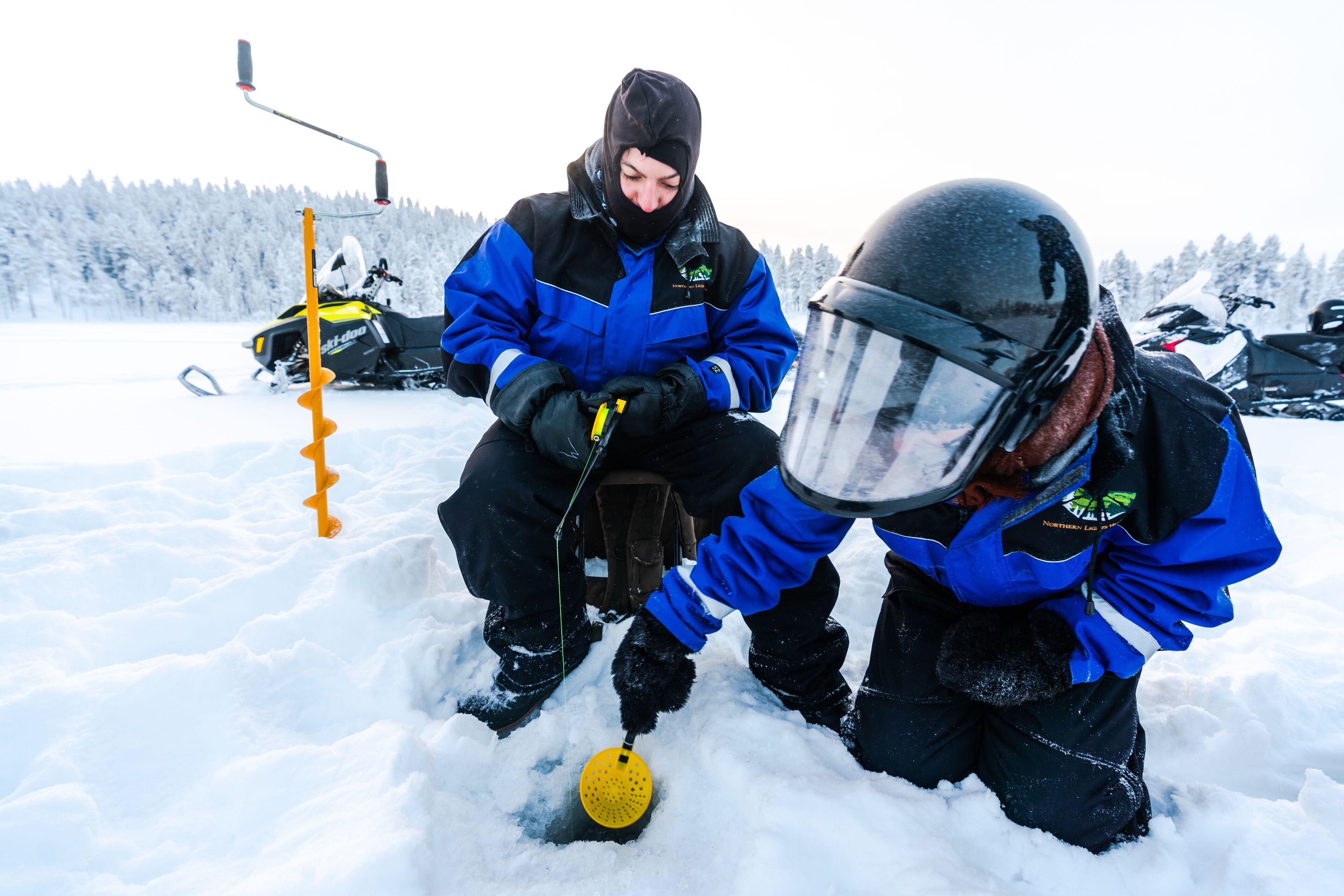 Two people in blue and black winter gear, one holding a yellow object, in a snowy outdoor setting with snowmobiles in the background.