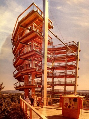 Una torre de observación de varios niveles, con una terraza de madera y barandillas de metal, rodeada de entorno natural.