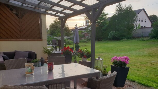 Een terras buiten met een tafel en stoelen, omringd door potplanten en uitzicht op een gazon.