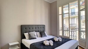 A bedroom with a large bed, a small side table, and a window with a view of buildings. - N&J - LILAS BUFFA - Central - Close sea (Niza)