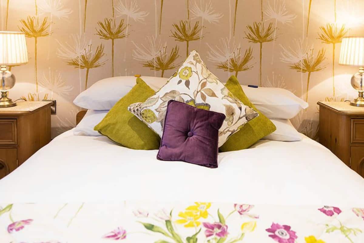 A neatly made bed with floral bedding, two bedside tables with lamps, and a floral-patterned wallpaper.