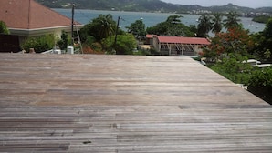 A wooden deck with a view of a coastal area, including a building with a red roof and a distant view of the sea and hills.