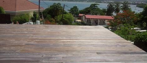 A wooden deck with a view of a coastal area, including a building with a red roof and a distant view of the sea and hills.