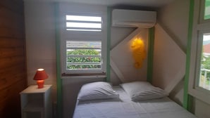 A small, cozy room with a bed, a nightstand, a window with a view of greenery, and an air conditioning unit.