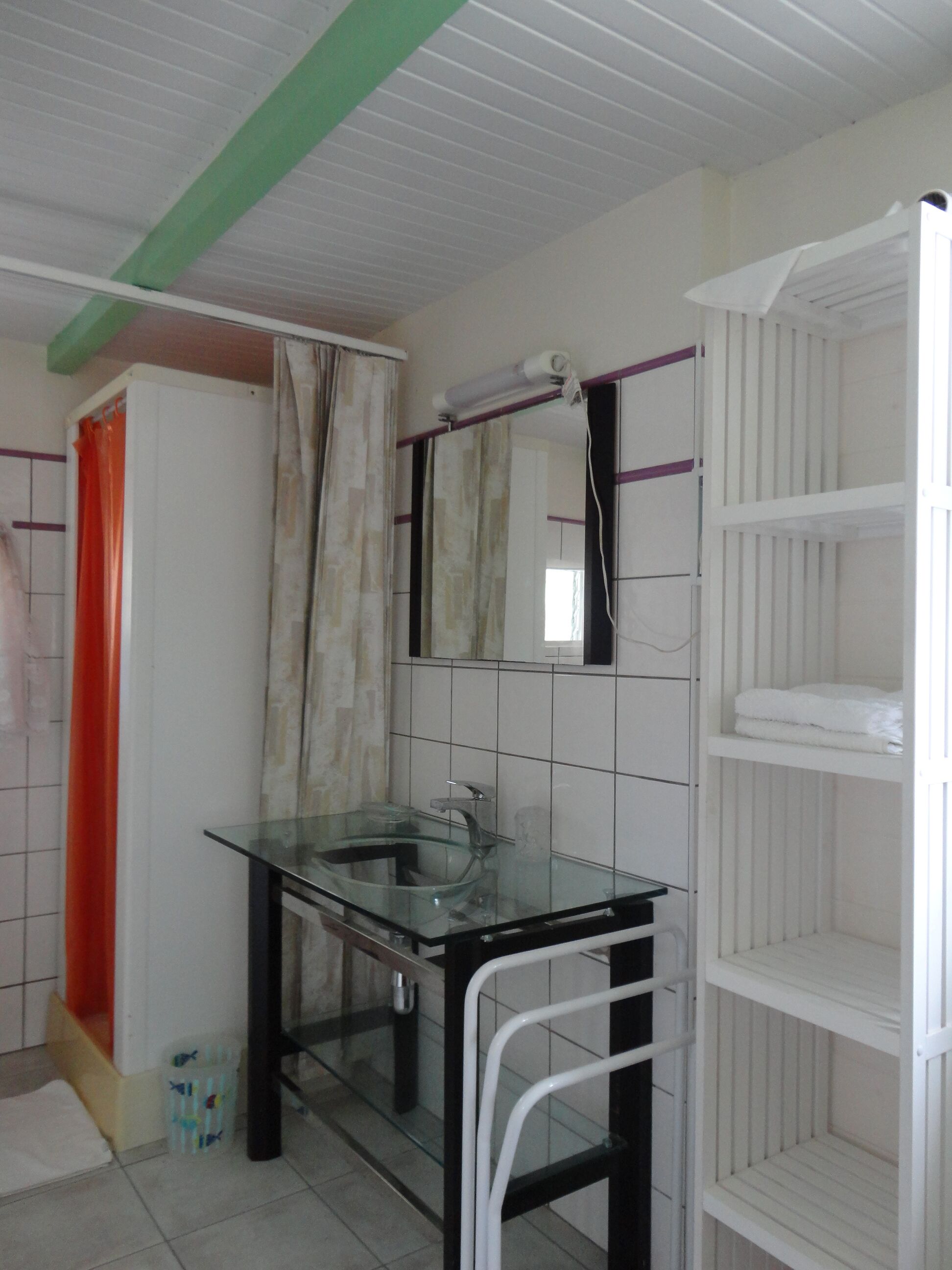 A bathroom with a glass sink, white tiled walls, a shower area, and a white shelf unit.