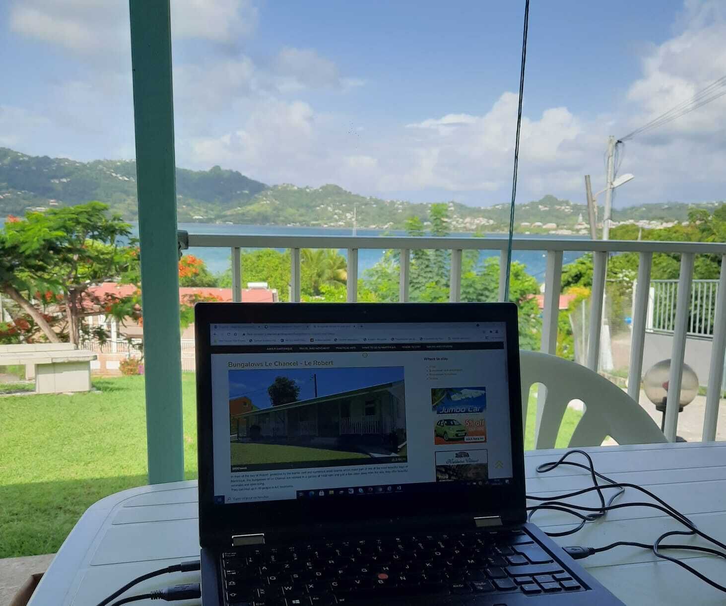 Ein Laptop auf einem Tisch, im Hintergrund eine grüne Landschaft und Gebäude.
