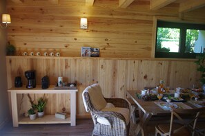 Un intérieur en bois avec une table à manger dressée pour le petit-déjeuner, une chaise en osier et une étagère ornée de plantes et d’ustensiles de cuisine.
