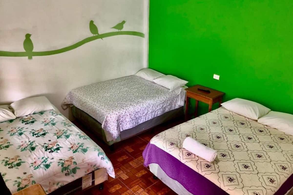 A room with three beds, each with a different patterned bedspread. There is a small wooden nightstand between the beds.