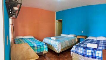 A room with three beds, each with a different color bedspread. There is a wooden bench and a small table.