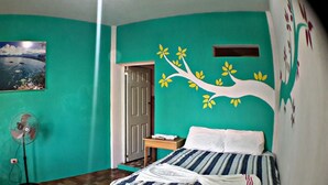Una habitación con paredes color turquesa, una cama con ropa de cama a rayas, un ventilador, una silla y un cuadro colgado en la pared de un paisaje.