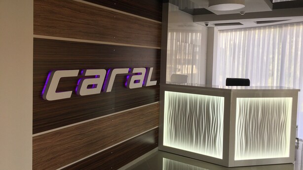 Bancada de recepção com design moderno, apresentando uma parede de painéis de madeira com a palavra "Caral" iluminada em roxo.
