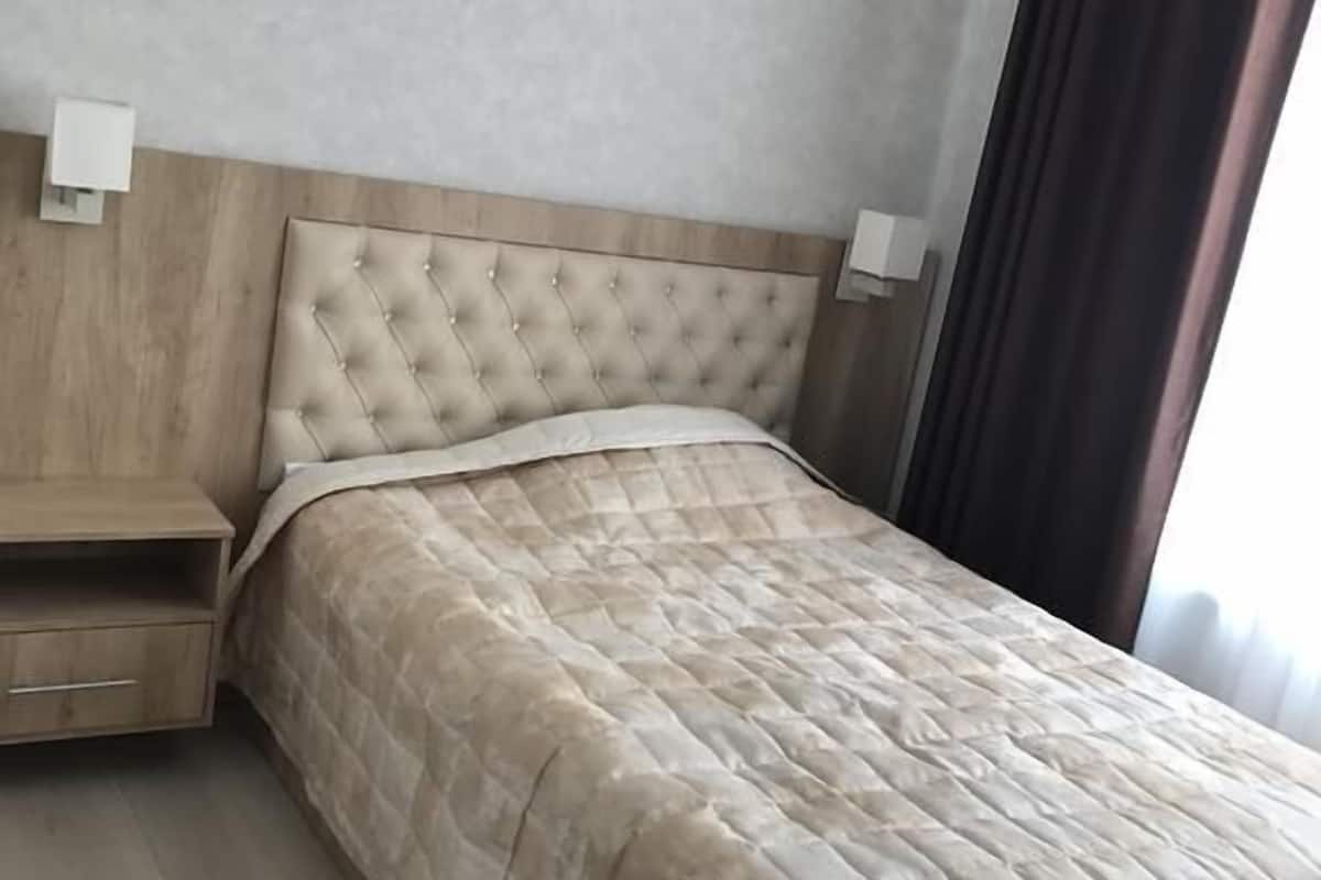 A bedroom with a tufted headboard, a wooden nightstand with a drawer, a bed with a beige quilt, and an air conditioning unit on the wall.