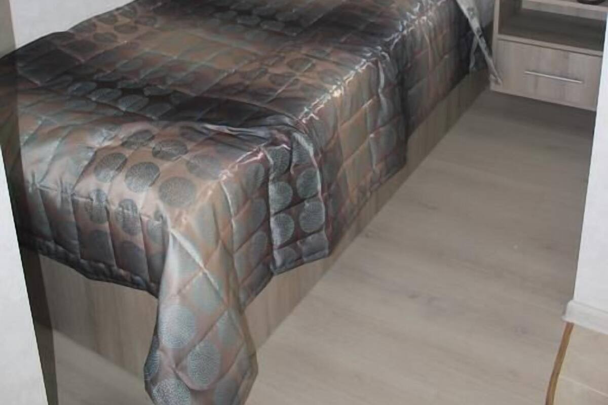 A single bed with a patterned bedspread, a bedside table with a bottle and tissues, and a wall-mounted TV.