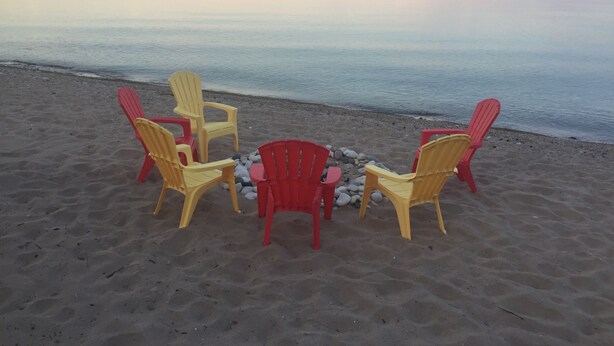 A beach with four Adirondack chairs arranged in a circle, a small table, and a pile of stones. - Lake Michigan Beach Home with Over 100’ of Private Sand Beach! Pets Welcome!  (Sturgeon Bay)
