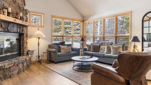 A cozy living room with a stone fireplace, large windows, a sofa set, and a leather armchair.