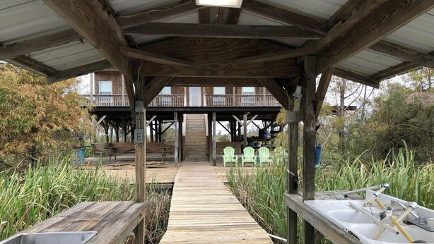 Un pontile in legno conduce a una zona riparata con posti a sedere all'aperto e vista su un edificio sullo sfondo.